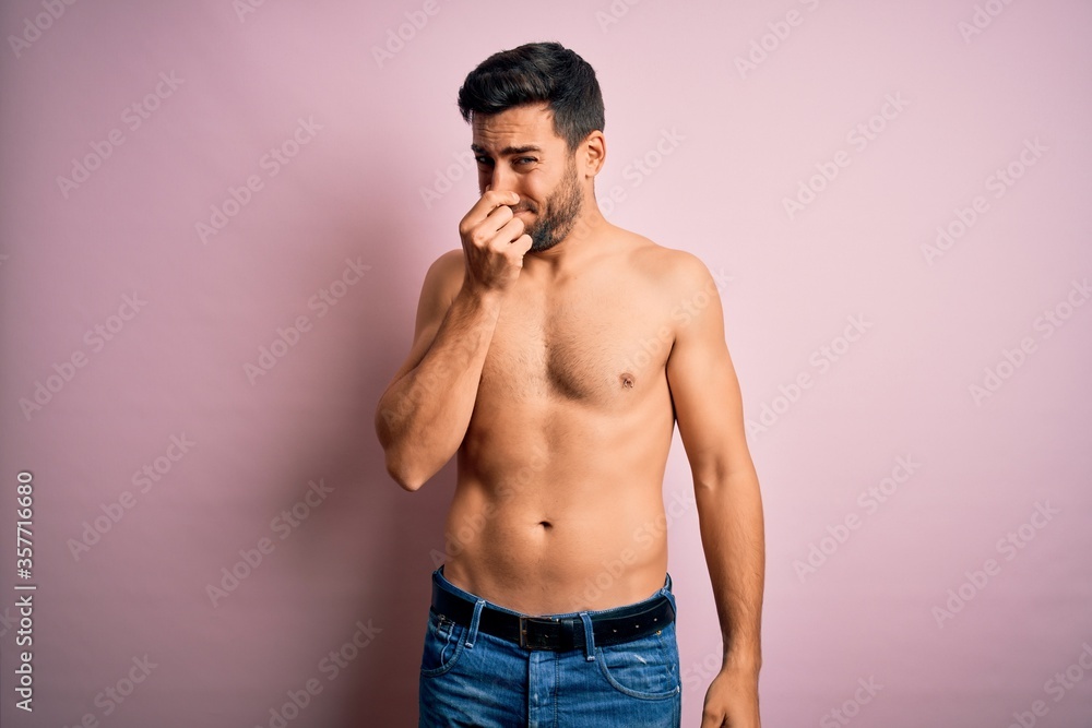 Young handsome strong man with beard shirtless standing over isolated pink background smelling something stinky and disgusting, intolerable smell, holding breath with fingers on nose. Bad smell