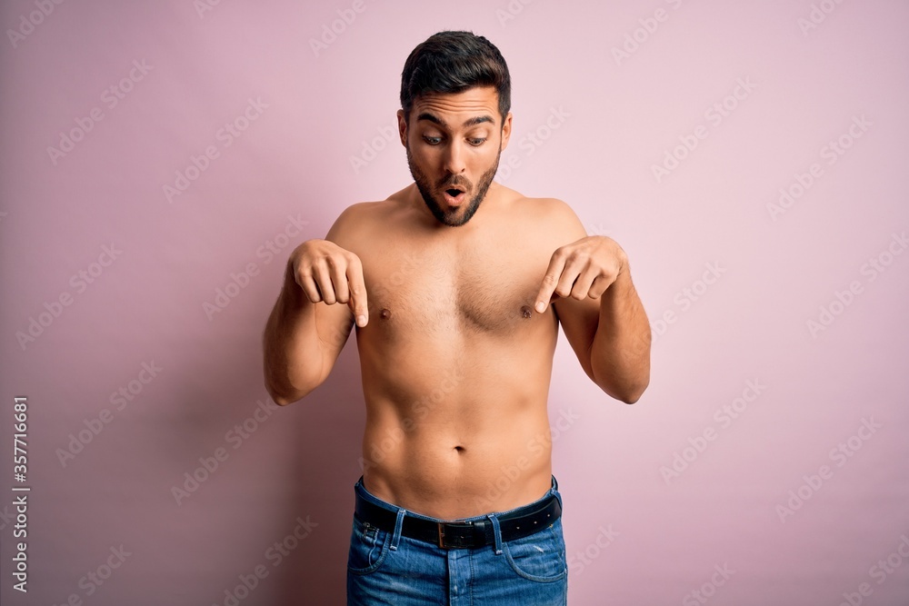 Fototapeta premium Young handsome strong man with beard shirtless standing over isolated pink background Pointing down with fingers showing advertisement, surprised face and open mouth
