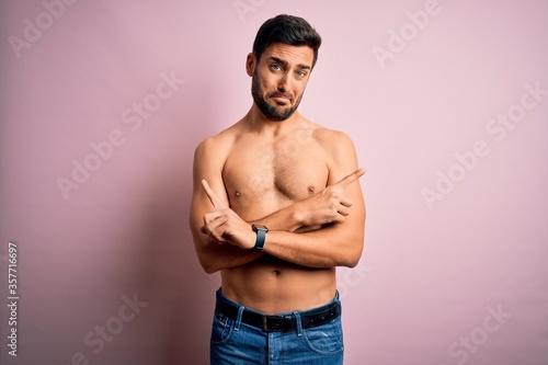Young handsome strong man with beard shirtless standing over isolated pink background Pointing to both sides with fingers, different direction disagree
