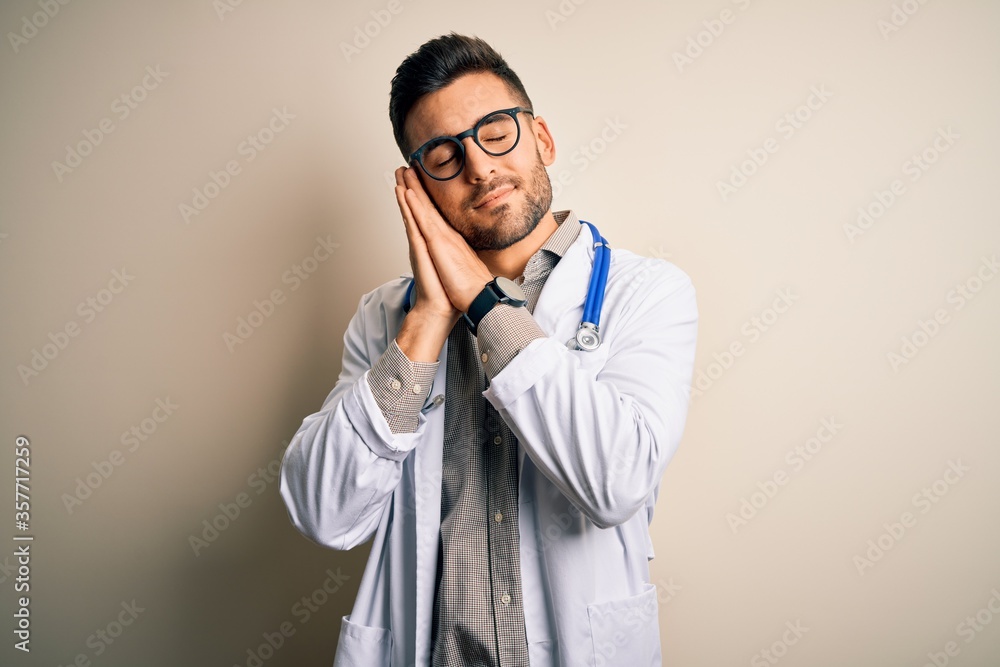 Young doctor man wearing glasses, medical white robe and stethoscope over isolated background sleeping tired dreaming and posing with hands together while smiling with closed eyes. Young doctor man wearing glasses, medical white robe and stethoscope over isolated background sleeping tired dreaming and posing with hands together while smiling with closed eyes.
