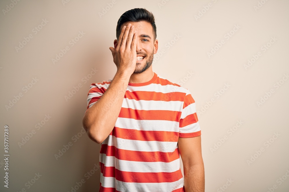 Young handsome man wearing casual striped t-shirt standing over isolated white background covering one eye with hand, confident smile on face and surprise emotion. Young handsome man wearing casual striped t-shirt standing over isolated white background covering one eye with hand, confident smile on face and surprise emotion.