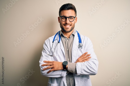 Young doctor man wearing glasses, medical white robe and stethoscope over isolated background happy face smiling with crossed arms looking at the camera. Positive person.