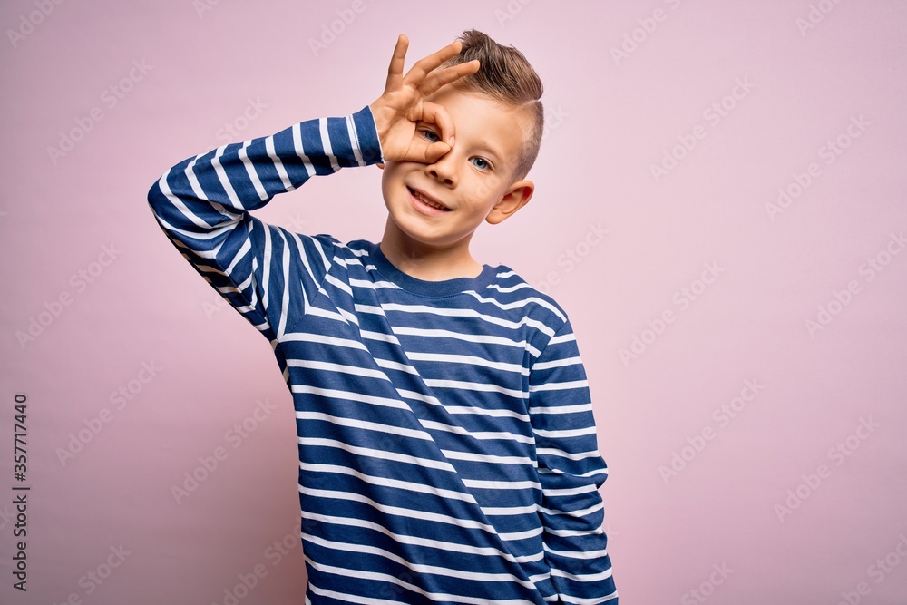 Young little caucasian kid with blue eyes wearing nautical striped shirt over pink background doing ok gesture with hand smiling, eye looking through fingers with happy face. Young little caucasian kid with blue eyes wearing nautical striped shirt over pink background doing ok gesture with hand smiling, eye looking through fingers with happy face.