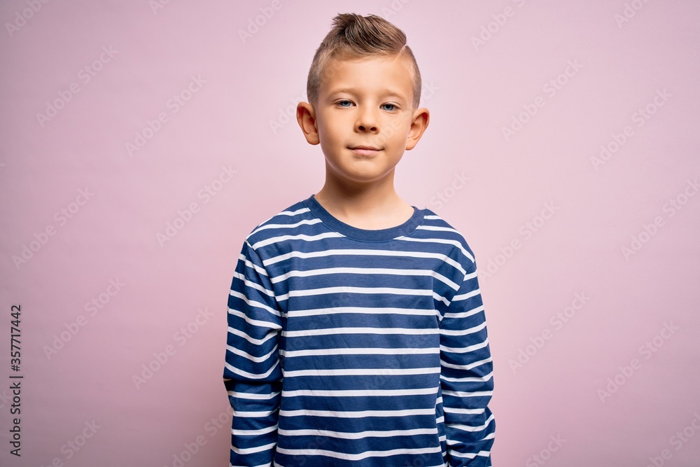 Young little caucasian kid with blue eyes wearing nautical striped shirt over pink background with serious expression on face. Simple and natural looking at the camera. Young little caucasian kid with blue eyes wearing nautical striped shirt over pink background with serious expression on face. Simple and natural looking at the camera.