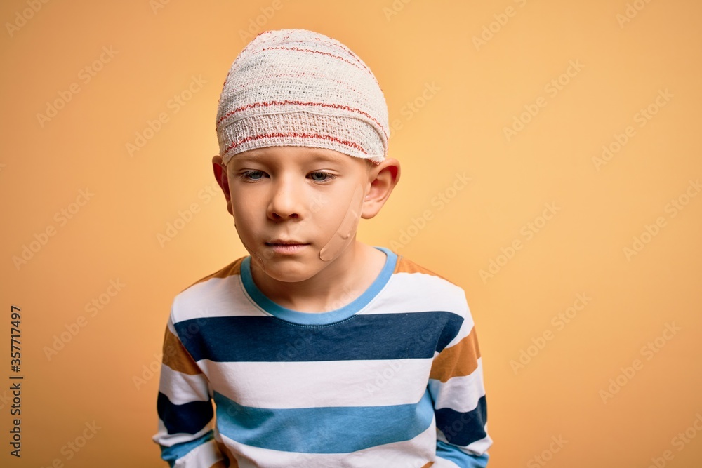 Young little caucasian kid injured wearing medical bandage on head over yellow background with hand on stomach because indigestion, painful illness feeling unwell. Ache concept.