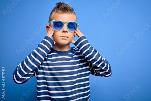 Young little caucasian kid with blue eyes standing wearing sunglasses over blue background suffering from headache desperate and stressed because pain and migraine. Hands on head.