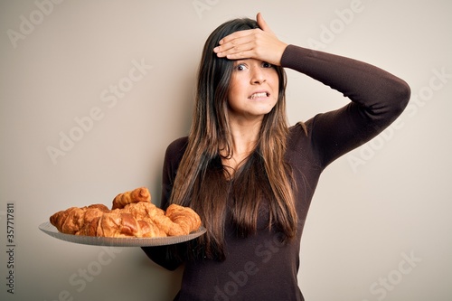 Young beautiful girl holding plate with sweet croissants for breakfast over white background stressed with hand on head, shocked with shame and surprise face, angry and frustrated. Fear and upset