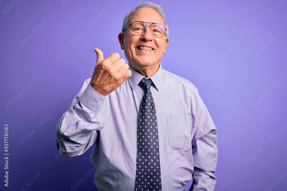 Grey haired senior business man wearing glasses standing over purple isolated background smiling with happy face looking and pointing to the side with thumb up. Grey haired senior business man wearing glasses standing over purple isolated background smiling with happy face looking and pointing to the side with thumb up.