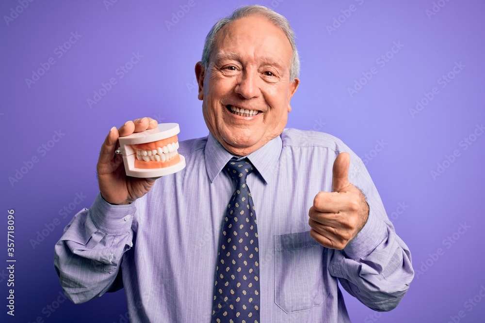 Grey haired senior man holding orthodontic prosthesis denture over purple background happy with big smile doing ok sign, thumb up with fingers, excellent sign Grey haired senior man holding orthodontic prosthesis denture over purple background happy with big smile doing ok sign, thumb up with fingers, excellent sign