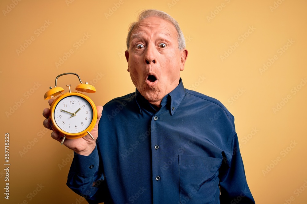 Senior grey haired man holding vintage alarm clock over yellow