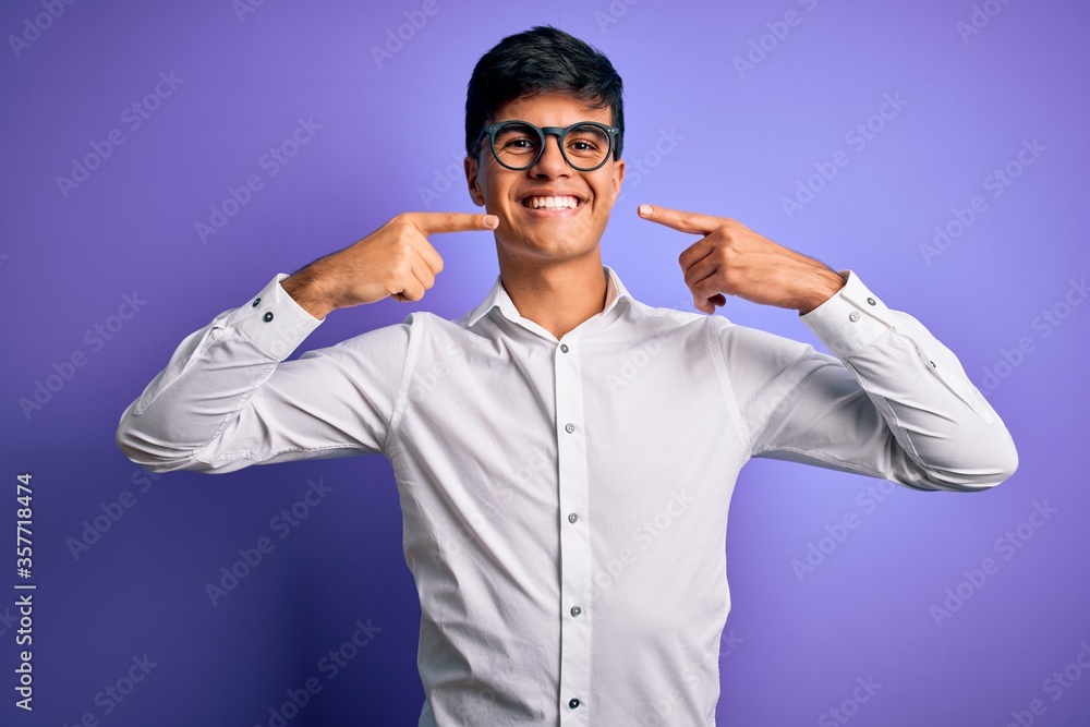 Young handsome business man wearing shirt and glasses over isolated purple background smiling cheerful showing and pointing with fingers teeth and mouth. Dental health concept. Young handsome business man wearing shirt and glasses over isolated purple background smiling cheerful showing and pointing with fingers teeth and mouth. Dental health concept.