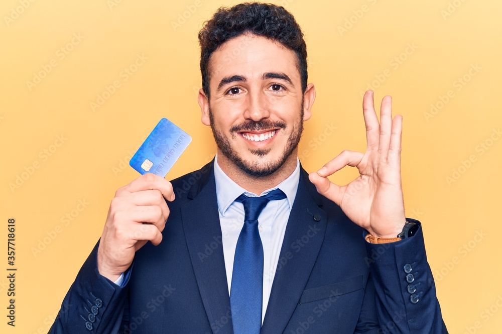 Young hispanic man wearing suit holding credit card doing ok sign with fingers, smiling friendly gesturing excellent symbol