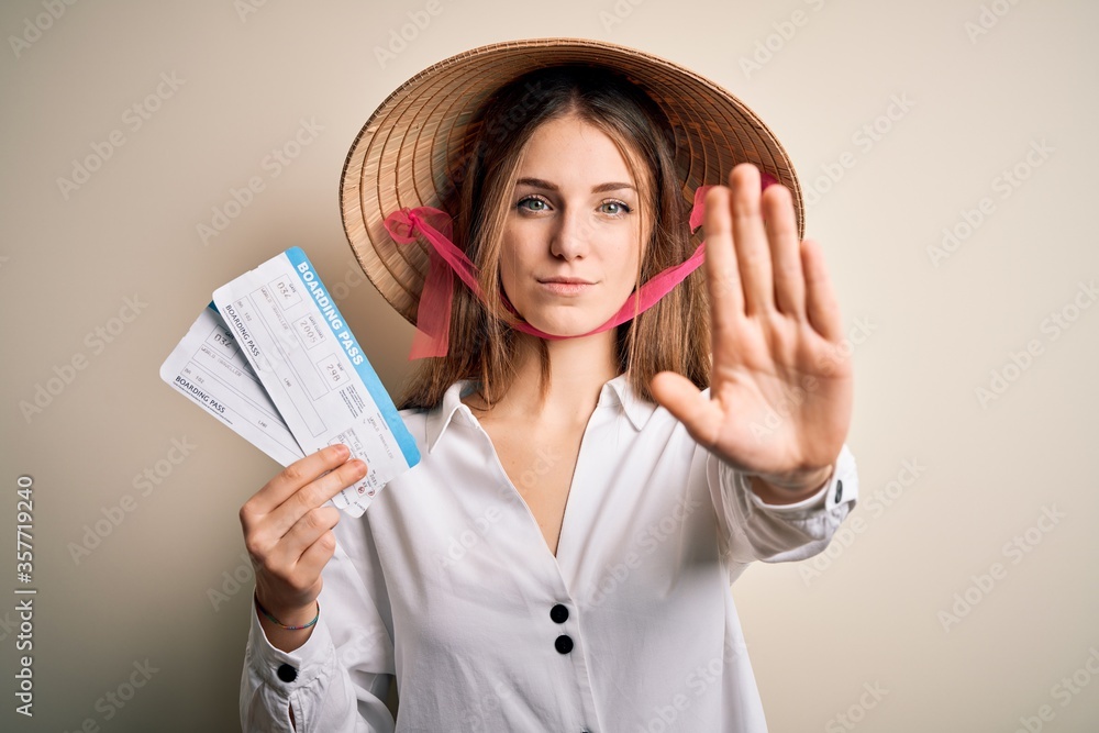 Young beautiful redhead tourist woman wearing asian traditional hat holding boarding pass with open hand doing stop sign with serious and confident expression, defense gesture Young beautiful redhead tourist woman wearing asian traditional hat holding boarding pass with open hand doing stop sign with serious and confident expression, defense gesture