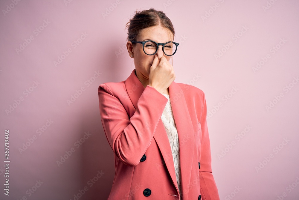 Young beautiful redhead woman wearing jacket and glasses over isolated pink background smelling something stinky and disgusting, intolerable smell, holding breath with fingers on nose. Bad smell