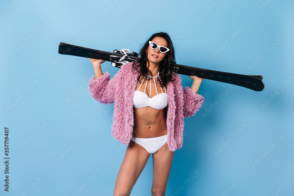 Confident stylish girl holding skis. Studio shot of adorable young woman in bikini and jacket isolated on blue background.