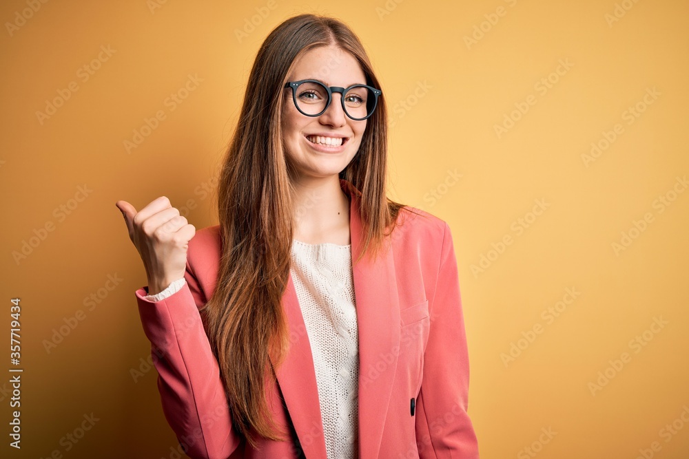 Young beautiful redhead woman wearing jacket and glasses over isolated yellow background smiling with happy face looking and pointing to the side with thumb up. Young beautiful redhead woman wearing jacket and glasses over isolated yellow background smiling with happy face looking and pointing to the side with thumb up.