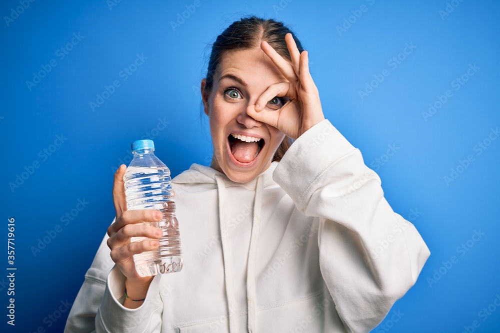 Young beautiful redhead woman doing sport drinking bottle of water over blue background with happy face smiling doing ok sign with hand on eye looking through fingers Young beautiful redhead woman doing sport drinking bottle of water over blue background with happy face smiling doing ok sign with hand on eye looking through fingers