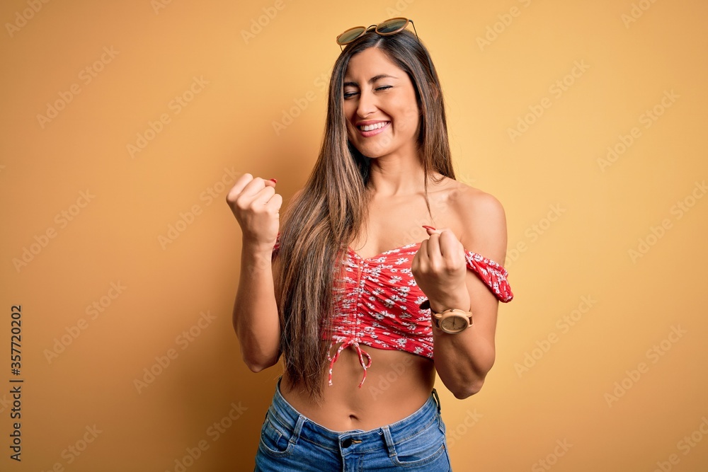 Young beautiful woman wearing summer style and sunglasses over yellow isolated background very happy and excited doing winner gesture with arms raised, smiling and screaming for success. Celebration Young beautiful woman wearing summer style and sunglasses over yellow isolated background very happy and excited doing winner gesture with arms raised, smiling and screaming for success. Celebration