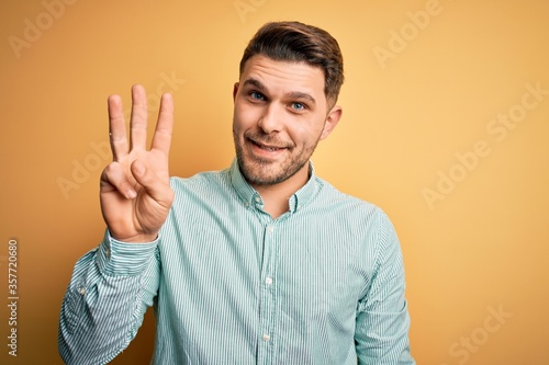 Young business man with blue eyes wearing elegant green shirt over yellow background showing and pointing up with fingers number three while smiling confident and happy.