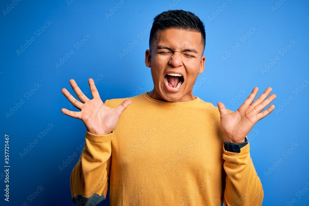 Young handsome latin man wearing yellow casual sweater over isolated blue background celebrating mad and crazy for success with arms raised and closed eyes screaming excited. Winner concept Young handsome latin man wearing yellow casual sweater over isolated blue background celebrating mad and crazy for success with arms raised and closed eyes screaming excited. Winner concept