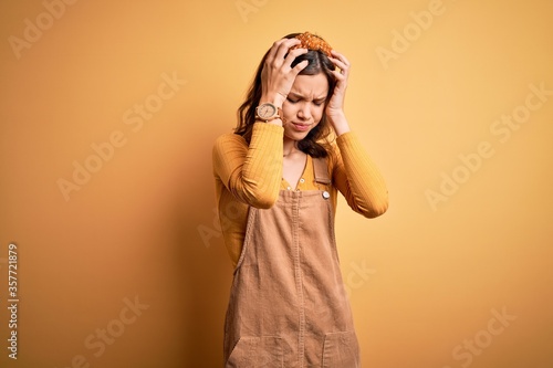 Young beautiful blonde girl wearing overall standing over yellow isolated background suffering from headache desperate and stressed because pain and migraine. Hands on head.