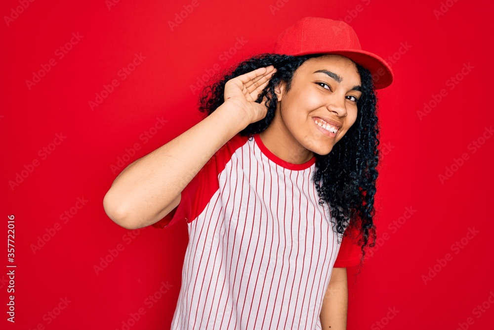 Young african american curly sportswoman wearing baseball cap and striped t-shirt smiling with hand over ear listening an hearing to rumor or gossip. Deafness concept. Young african american curly sportswoman wearing baseball cap and striped t-shirt smiling with hand over ear listening an hearing to rumor or gossip. Deafness concept.
