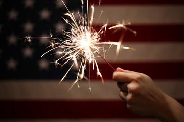 Hand holding lit sparkler in front of the American Flag for 4th of July celebration