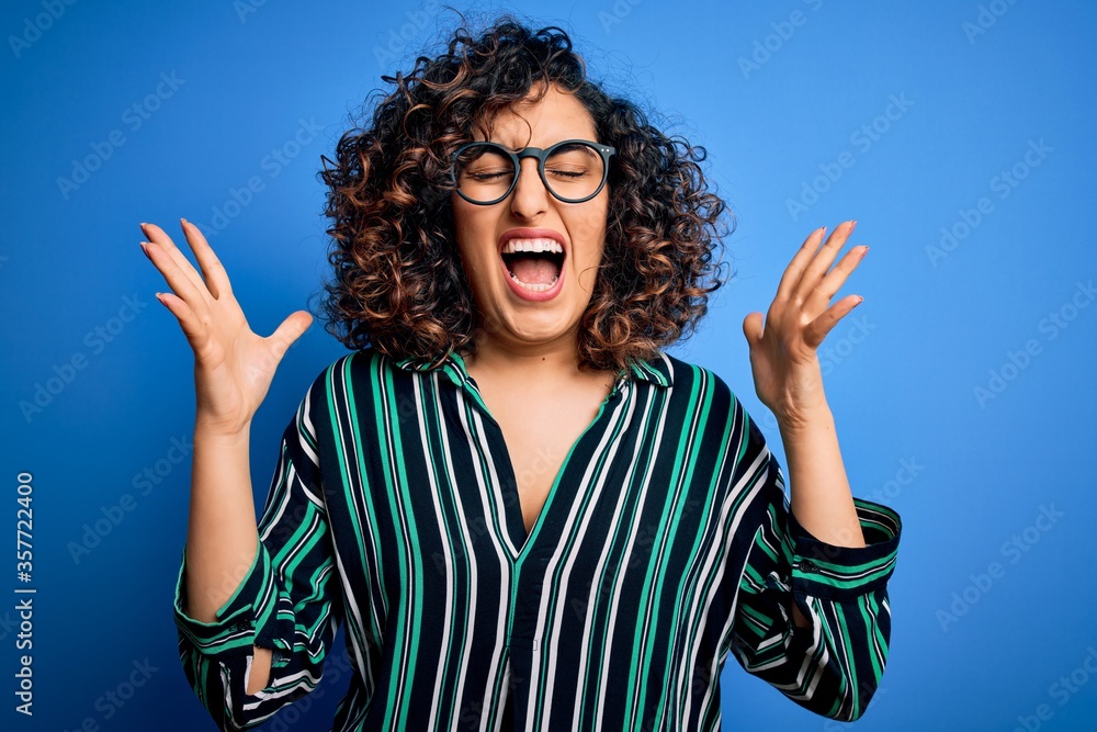 Young beautiful curly arab woman wearing striped shirt and glasses over blue background celebrating mad and crazy for success with arms raised and closed eyes screaming excited. Winner concept Young beautiful curly arab woman wearing striped shirt and glasses over blue background celebrating mad and crazy for success with arms raised and closed eyes screaming excited. Winner concept