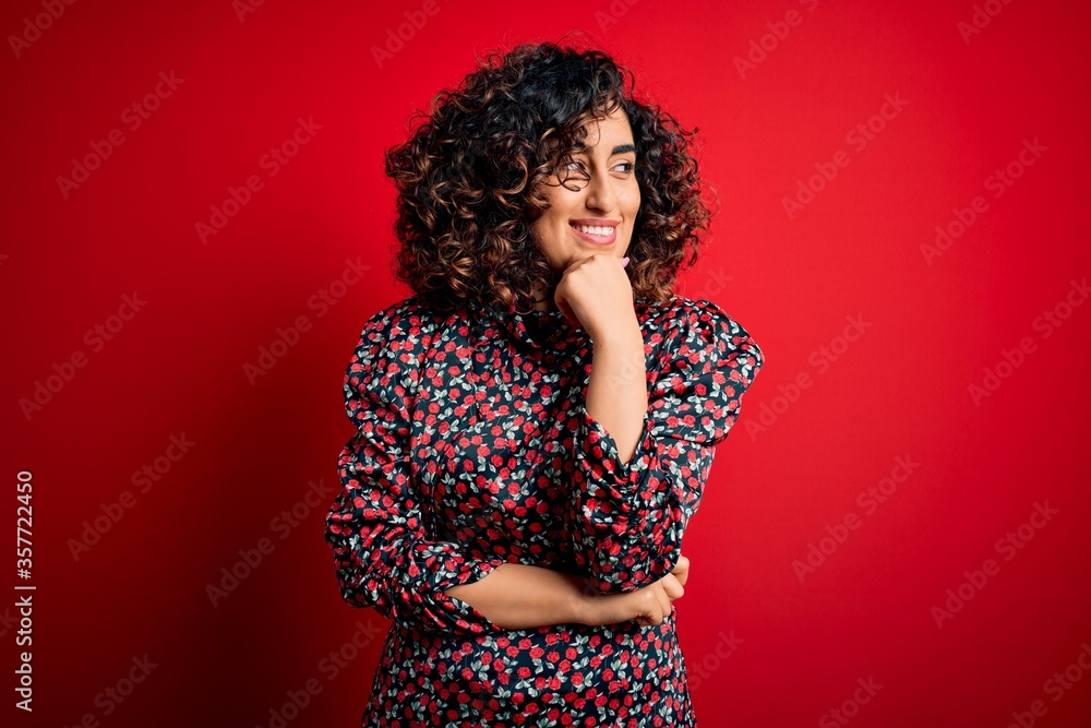 Young beautiful curly arab woman wearing casual floral dress standing over red background with hand on chin thinking about question, pensive expression. Smiling with thoughtful face. Doubt concept. Young beautiful curly arab woman wearing casual floral dress standing over red background with hand on chin thinking about question, pensive expression. Smiling with thoughtful face. Doubt concept.