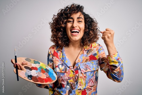 Young beautiful curly arab artist woman painting using brush and palette with colors screaming proud and celebrating victory and success very excited, cheering emotion photo