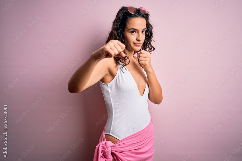Beautiful woman with curly hair on vacation wearing white swimsuit over pink background Punching fist to fight, aggressive and angry attack, threat and violence Beautiful woman with curly hair on vacation wearing white swimsuit over pink background Punching fist to fight, aggressive and angry attack, threat and violence