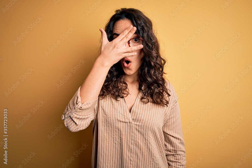 Beautiful woman with curly hair wearing striped shirt and glasses over yellow background peeking in shock covering face and eyes with hand, looking through fingers with embarrassed expression. Beautiful woman with curly hair wearing striped shirt and glasses over yellow background peeking in shock covering face and eyes with hand, looking through fingers with embarrassed expression.