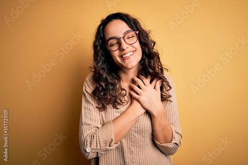Beautiful woman with curly hair wearing striped shirt and glasses over yellow background smiling with hands on chest with closed eyes and grateful gesture on face. Health concept.