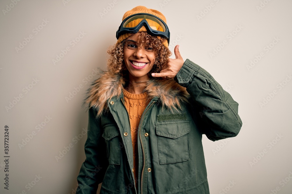 Young african american skier woman with curly hair wearing snow sportswear and ski goggles smiling doing phone gesture with hand and fingers like talking on the telephone. Communicating concepts. Young african american skier woman with curly hair wearing snow sportswear and ski goggles smiling doing phone gesture with hand and fingers like talking on the telephone. Communicating concepts.