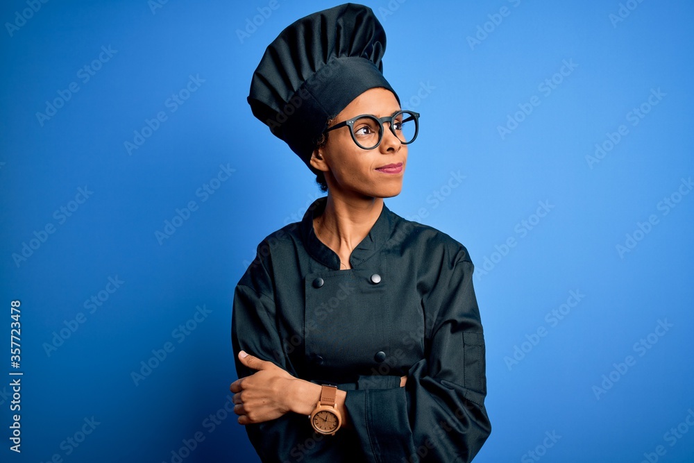 Young african american chef woman wearing cooker uniform and hat over blue background looking to the side with arms crossed convinced and confident Young african american chef woman wearing cooker uniform and hat over blue background looking to the side with arms crossed convinced and confident
