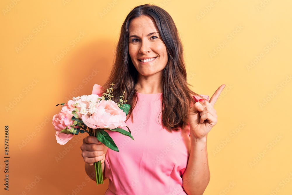 Young beautiful brunette woman holding bouquet of pink flowers over yellow background smiling happy pointing with hand and finger to the side Young beautiful brunette woman holding bouquet of pink flowers over yellow background smiling happy pointing with hand and finger to the side