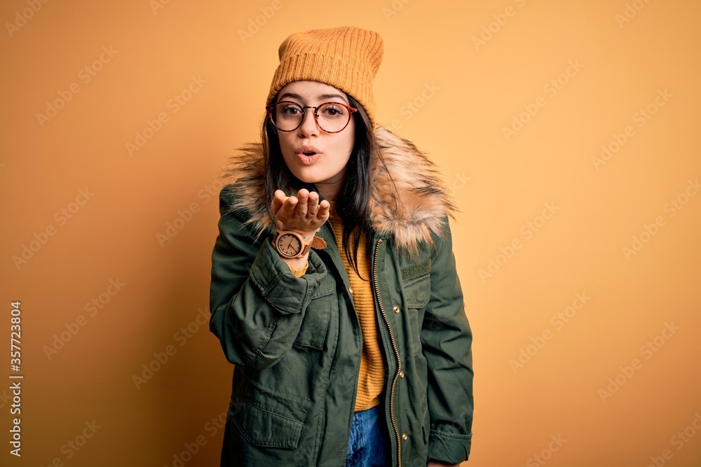 Young brunette woman wearing glasses and winter coat with hat over yellow isolated background looking at the camera blowing a kiss with hand on air being lovely and sexy. Love expression. Young brunette woman wearing glasses and winter coat with hat over yellow isolated background looking at the camera blowing a kiss with hand on air being lovely and sexy. Love expression.