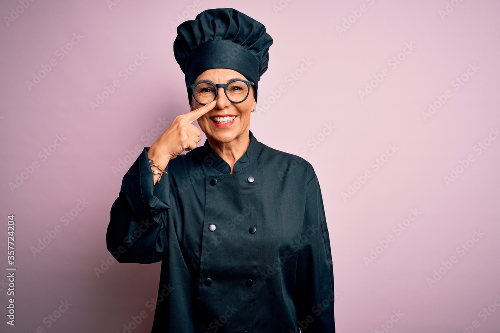 Middle age brunette chef woman wearing cooker uniform and hat over isolated pink background Pointing with hand finger to face and nose, smiling cheerful. Beauty concept Middle age brunette chef woman wearing cooker uniform and hat over isolated pink background Pointing with hand finger to face and nose, smiling cheerful. Beauty concept