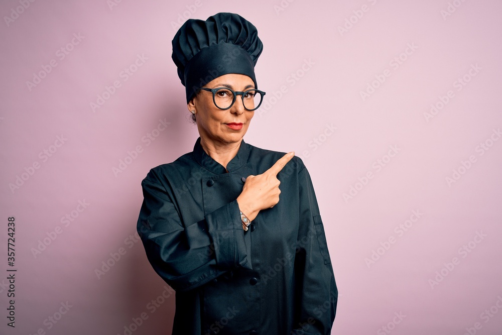 Middle age brunette chef woman wearing cooker uniform and hat over isolated pink background Pointing with hand finger to the side showing advertisement, serious and calm face Middle age brunette chef woman wearing cooker uniform and hat over isolated pink background Pointing with hand finger to the side showing advertisement, serious and calm face