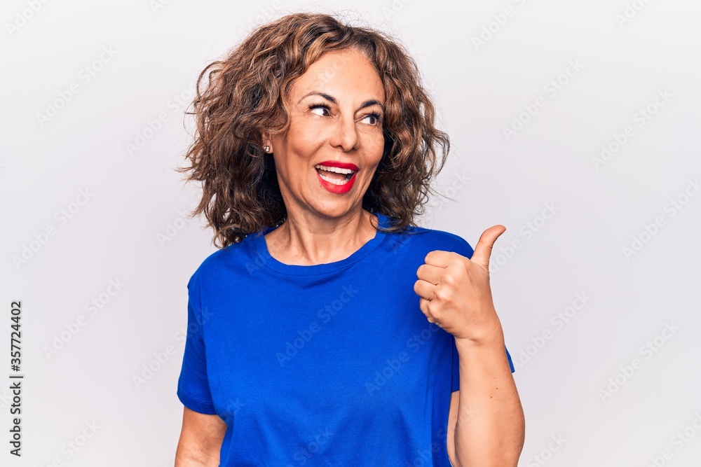 Middle age beautiful brunette woman wearing blue t-shirt standing over white background pointing thumb up to the side smiling happy with open mouth Middle age beautiful brunette woman wearing blue t-shirt standing over white background pointing thumb up to the side smiling happy with open mouth