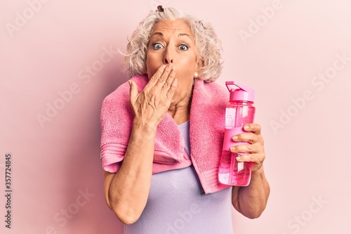 Senior grey-haired woman wearing sportswear and towel drinking bottle of water covering mouth with hand, shocked and afraid for mistake. surprised expression