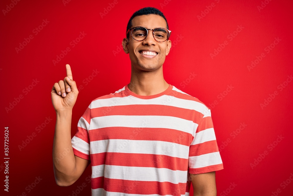 Fototapeta premium Young handsome african american man wearing casual striped t-shirt and glasses with a big smile on face, pointing with hand finger to the side looking at the camera.