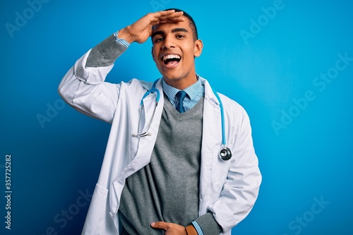 Handsome african american doctor man wearing coat and stethoscope over blue background very happy and smiling looking far away with hand over head. Searching concept.