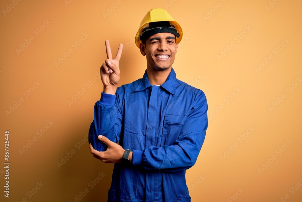 Young handsome african american worker man wearing blue uniform and security helmet smiling with happy face winking at the camera doing victory sign with fingers. Number two.