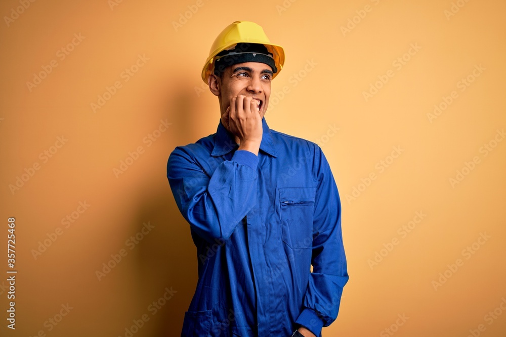 Young handsome african american worker man wearing blue uniform and security helmet looking stressed and nervous with hands on mouth biting nails. Anxiety problem.