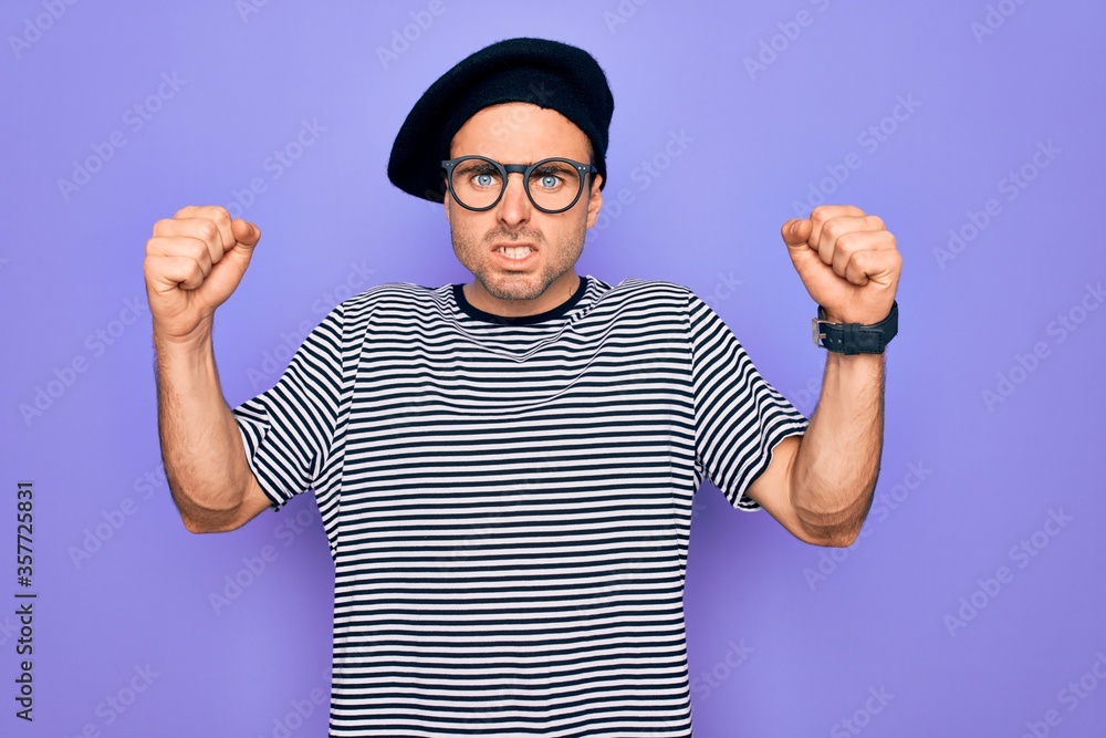 Fototapeta premium Handsome man with blue eyes wearing striped t-shirt and french beret over purple background angry and mad raising fists frustrated and furious while shouting with anger. Rage and aggressive concept.