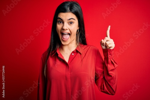 Young beautiful brunette woman wearing casual shirt standing over red background pointing finger up with successful idea. Exited and happy. Number one.