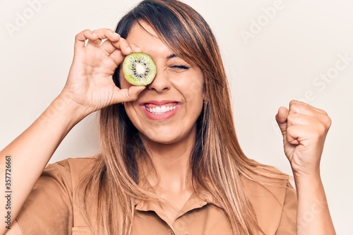 Young beautiful woman holding kiwi over eye screaming proud, celebrating victory and success very excited with raised arm