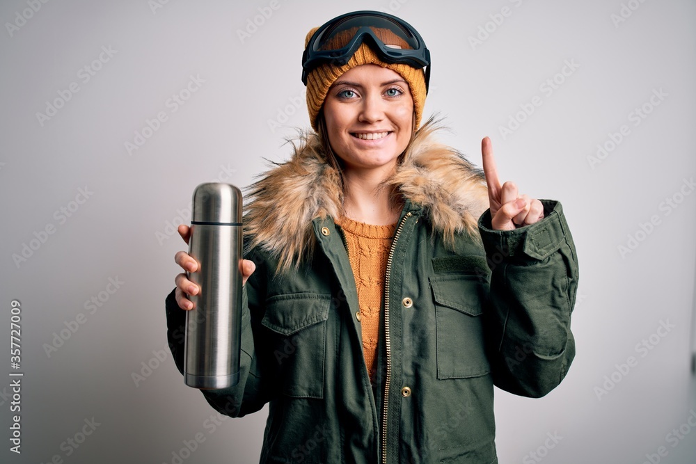 Young beautiful skier woman with blue eyes wearing ski goggles drinking thermo with coffee surprised with an idea or question pointing finger with happy face, number one Young beautiful skier woman with blue eyes wearing ski goggles drinking thermo with coffee surprised with an idea or question pointing finger with happy face, number one
