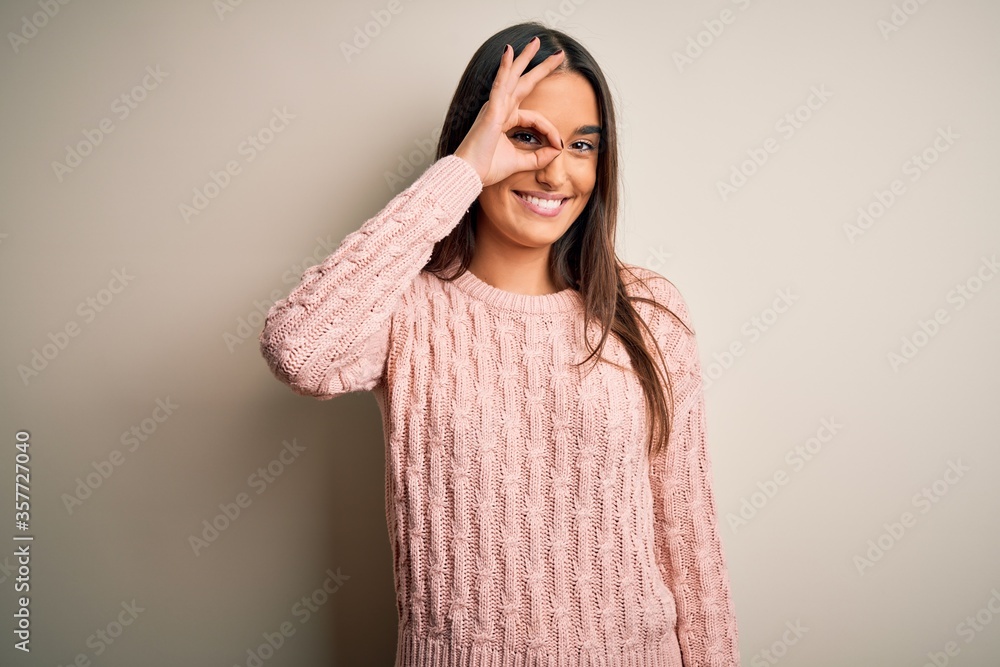 Young beautiful brunette woman wearing casual sweater over isolated white background doing ok gesture with hand smiling, eye looking through fingers with happy face. Young beautiful brunette woman wearing casual sweater over isolated white background doing ok gesture with hand smiling, eye looking through fingers with happy face.